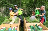 Zwei Erwachsene und zwei Kinder gießen mit Gießkannen Wasser in einen Miniatur-Wald